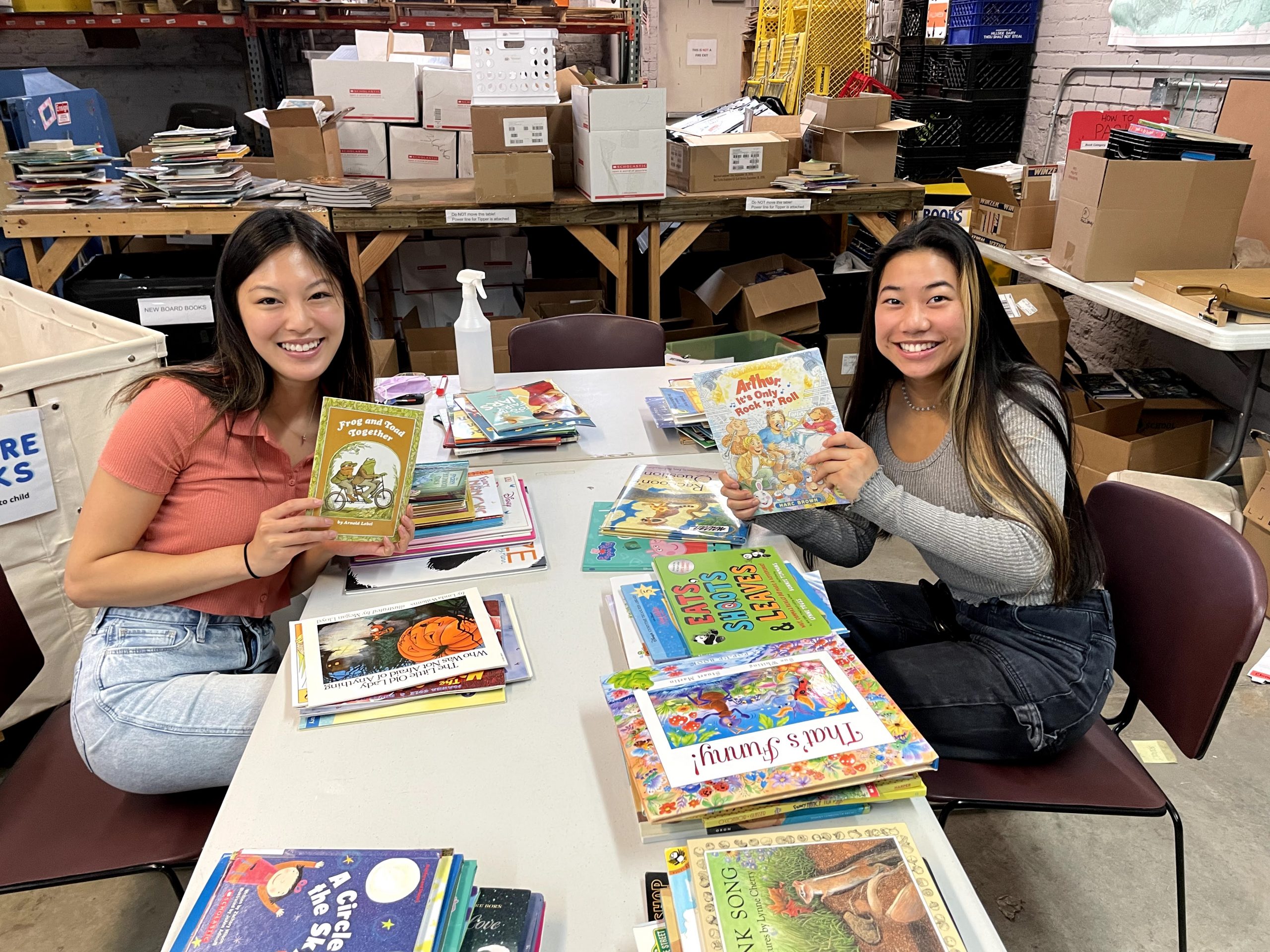 IMG_5905 Two smiling young women seated at a table of books at the Cleveland Kids' Book Bank warehouse