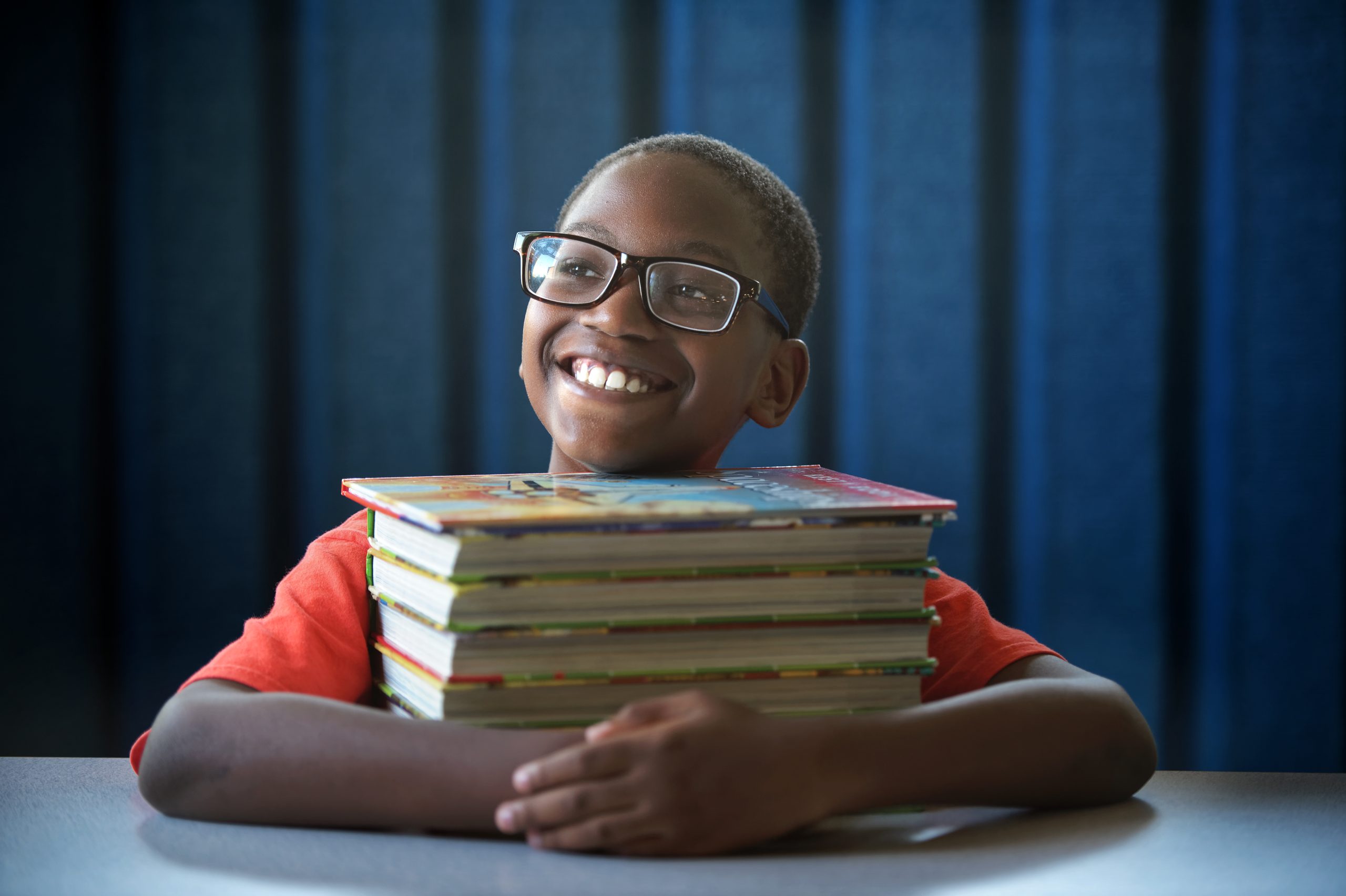 boy with glasses circling his arms around a stack of books on a table and smiling off the right
