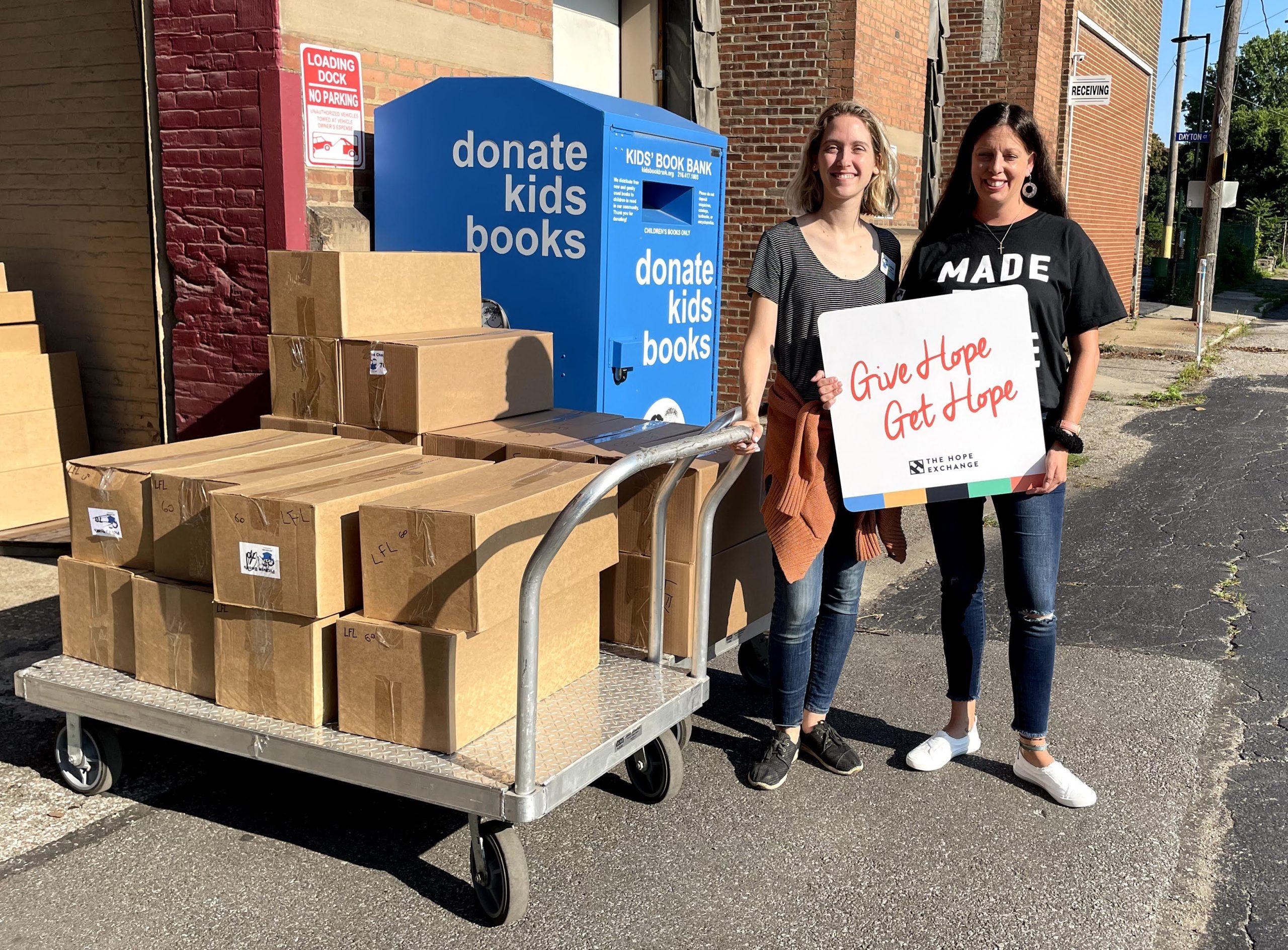 two women standing outside next to a cart full of boxes