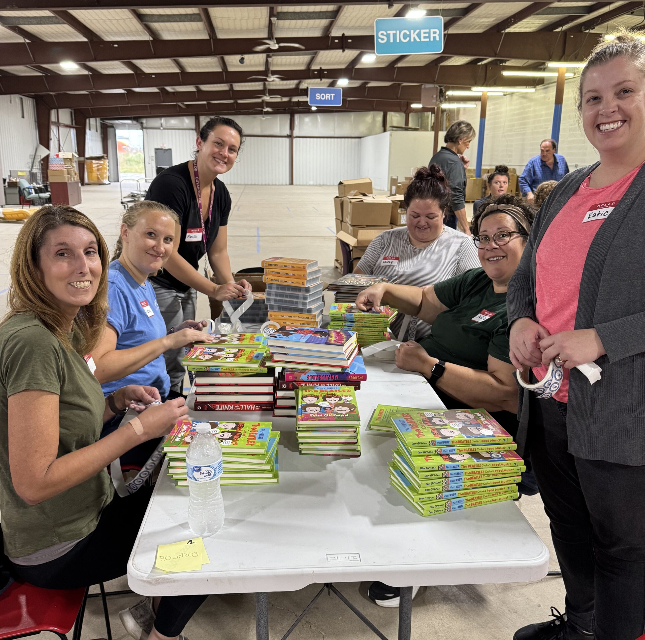 woman stands behind a table smiling and holding a tape dispenser